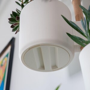 Puede incluir: Una maceta colgante cil&iacute;ndrica blanca con una base redondeada. La maceta est&aacute; dise&ntilde;ada para contener una planta peque&ntilde;a, con una suculenta visible en la parte superior. La maceta est&aacute; suspendida de un brazo de madera.