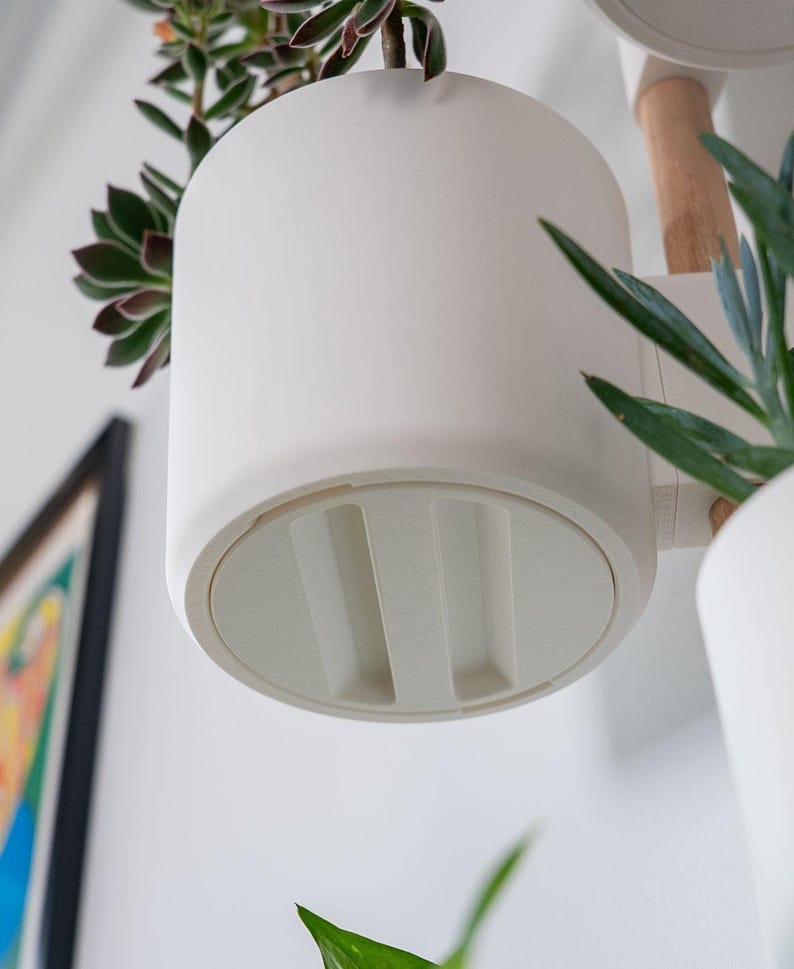 Puede incluir: Una maceta colgante cil&iacute;ndrica blanca con una base redondeada. La maceta est&aacute; dise&ntilde;ada para contener una planta peque&ntilde;a, con una suculenta visible en la parte superior. La maceta est&aacute; suspendida de un brazo de madera.