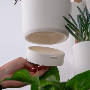 Puede incluir: Jardinera colgante de cer&aacute;mica blanca con base extra&iacute;ble, dise&ntilde;ada para plantas de interior. La jardinera es cil&iacute;ndrica y est&aacute; suspendida de un soporte de madera. Se ven plantas verdes y follaje, lo que sugiere una est&eacute;tica moderna y minimalista.