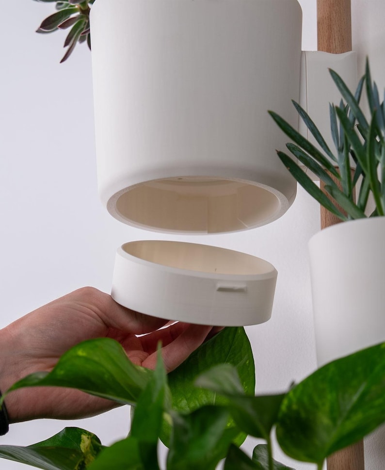 Puede incluir: Jardinera colgante de cer&aacute;mica blanca con base extra&iacute;ble, dise&ntilde;ada para plantas de interior. La jardinera es cil&iacute;ndrica y est&aacute; suspendida de un soporte de madera. Se ven plantas verdes y follaje, lo que sugiere una est&eacute;tica moderna y minimalista.