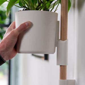 Puede incluir: Una maceta blanca montada en la pared con un soporte de madera. La maceta contiene una planta de hojas verdes. El soporte de madera est&aacute; fijado a la pared con soportes blancos. La maceta est&aacute; sujeta por una mano.