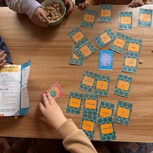 May include: A wooden table displays a children's game with cards featuring Arabic script and colorful patterns. A bowl of cereal and a game instruction booklet are also visible. Children's hands are interacting with the game.