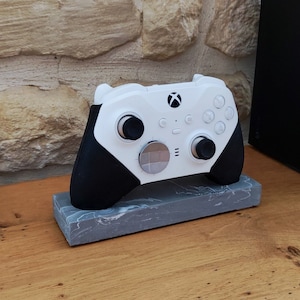 Op de afbeelding: Een witte en zwarte Xbox-controller met een grijze marmeren standaard. De controller heeft een witte voorplaat met zwarte knoppen en een zwarte achterkant.