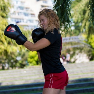Puede incluir: Una mujer con guantes de boxeo negros y pantalones cortos rojos está practicando un jab en un parque.