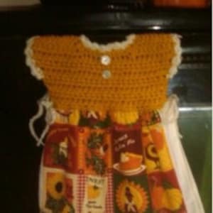 May include: A crocheted dish towel holder in the shape of a dress. The dress is yellow with white trim and buttons. The bottom of the dress is a white tea towel with a colorful print of fruits and vegetables.