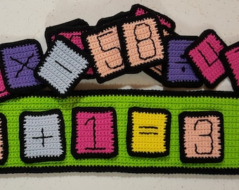 May include: A green felt board with a black border and a red apple. The board has a math equation written on it: 2 + 1 = 3. There are colorful crocheted squares with numbers and symbols on top of the board.