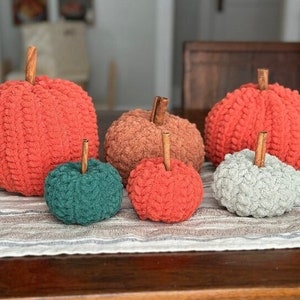Puede incluir: Un conjunto de seis calabazas de ganchillo en varios tamaños y colores, incluyendo naranja, verde y gris. Las calabazas están dispuestas sobre un camino de mesa a rayas.