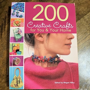 May include: A softcover book titled 200 Creative Crafts for You & Your Home, edited by Megan Hiller. The cover features a large magenta background with white and yellow text, and a side panel displaying six small craft project photographs.