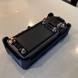 May include: A black audio recording device with a screen and multiple silver knobs. The device is labeled "SHURE" and is housed in a black protective case. The setup is on a light-colored surface.