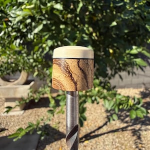 May include: A wooden bottle stopper with a light-colored top and a cylindrical body featuring a unique wood grain pattern in shades of brown and tan. The stopper is mounted on a silver metal rod, showcasing a handcrafted design.
