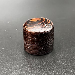 May include: A cylindrical wooden knob with a dark brown finish and a glossy top. The top features a lighter brown, swirling wood grain pattern. The knob is likely for furniture or a similar application.