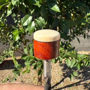 May include: A wooden mallet with a light-colored top and a reddish-brown body. The body has a textured surface. The mallet is attached to a metal shaft.