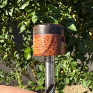 Puede incluir: Una broca con mango de madera. El mango es un cilindro con una parte superior marrón oscuro y un cuerpo marrón claro texturizado. Un pequeño agujero es visible en el lateral del mango. La broca es plateada.