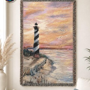 May include: A rectangular woven throw blanket featuring a lighthouse with black and white stripes on a sandy shore at sunset. The sky is filled with pink, orange, and purple hues, and two birds are flying. The blanket has a fringed edge.