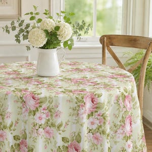 May include: A round tablecloth featuring a light green and white vertical striped pattern with large pink rose clusters and green leaves. A white ceramic pitcher holds a bouquet of white hydrangea flowers and green eucalyptus stems.