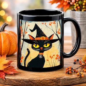 May include: A black ceramic mug featuring a graphic illustration of a black cat wearing a witch hat with a yellow buckle. The cat has bright yellow eyes and an orange collar with a small circular charm, set against a light background with trees.