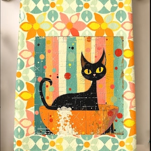 May include: A rectangular hand towel featuring a repeating pattern of orange, yellow, and teal flowers. The center displays a black cat with yellow eyes sitting in an orange bowl filled with white bubbles against a vertical striped background.
