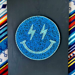 May include: A string art piece featuring a blue smiley face with lightning bolt eyes on a black wooden board. The smiley face is made of blue string, with light blue accents. The board is rectangular.
