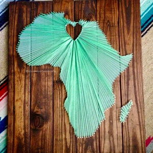 May include: A green string art piece of the continent of Africa with a heart shape in the center. The art is mounted on a wooden board.