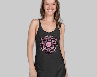 May include: A dark heather gray racerback tank top featuring a central pink graphic. The design consists of a solid pink circle containing a black infinity-style symbol, surrounded by radiating pink lines that create a sunburst effect.
