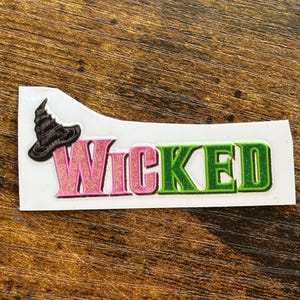 Może przedstawiać: Naklejka z napisem "Wicked" w kolorze r&oacute;żowym i zielonym, z czarnym kapeluszem czarownicy. Naklejka znajduje się na białym tle, na drewnianej powierzchni.