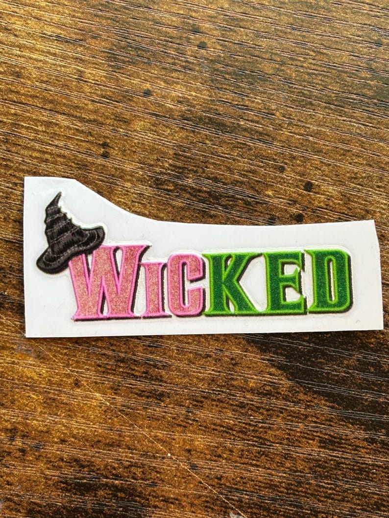 Może przedstawiać: Naklejka z napisem "Wicked" w kolorze r&oacute;żowym i zielonym, z czarnym kapeluszem czarownicy. Naklejka znajduje się na białym tle, na drewnianej powierzchni.