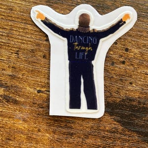 Może przedstawiać: Naklejka przedstawiająca postać w ciemnoniebieskim stroju z wyciągniętymi ramionami, ozdobiona złotymi detalami. Tekst "Dancing Through Life" jest napisany złotym pismem. Naklejka ma białą obw&oacute;dkę.
