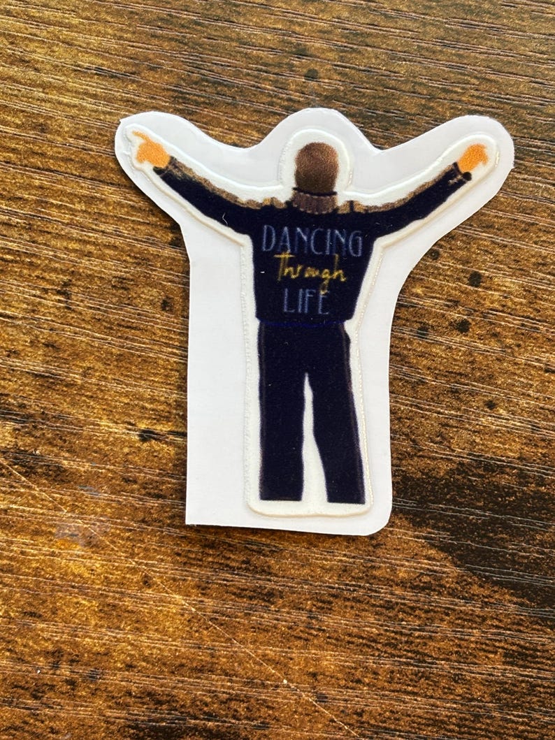 Może przedstawiać: Naklejka przedstawiająca postać w ciemnoniebieskim stroju z wyciągniętymi ramionami, ozdobiona złotymi detalami. Tekst "Dancing Through Life" jest napisany złotym pismem. Naklejka ma białą obw&oacute;dkę.