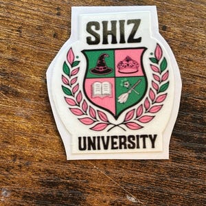 Może przedstawiać: Biała naklejka z czarnym napisem "SHIZ UNIVERSITY". Naklejka przedstawia herb w zielono-r&oacute;żowej kolorystyce, zawierający kapelusz czarownicy, koronę, książkę i miotłę.