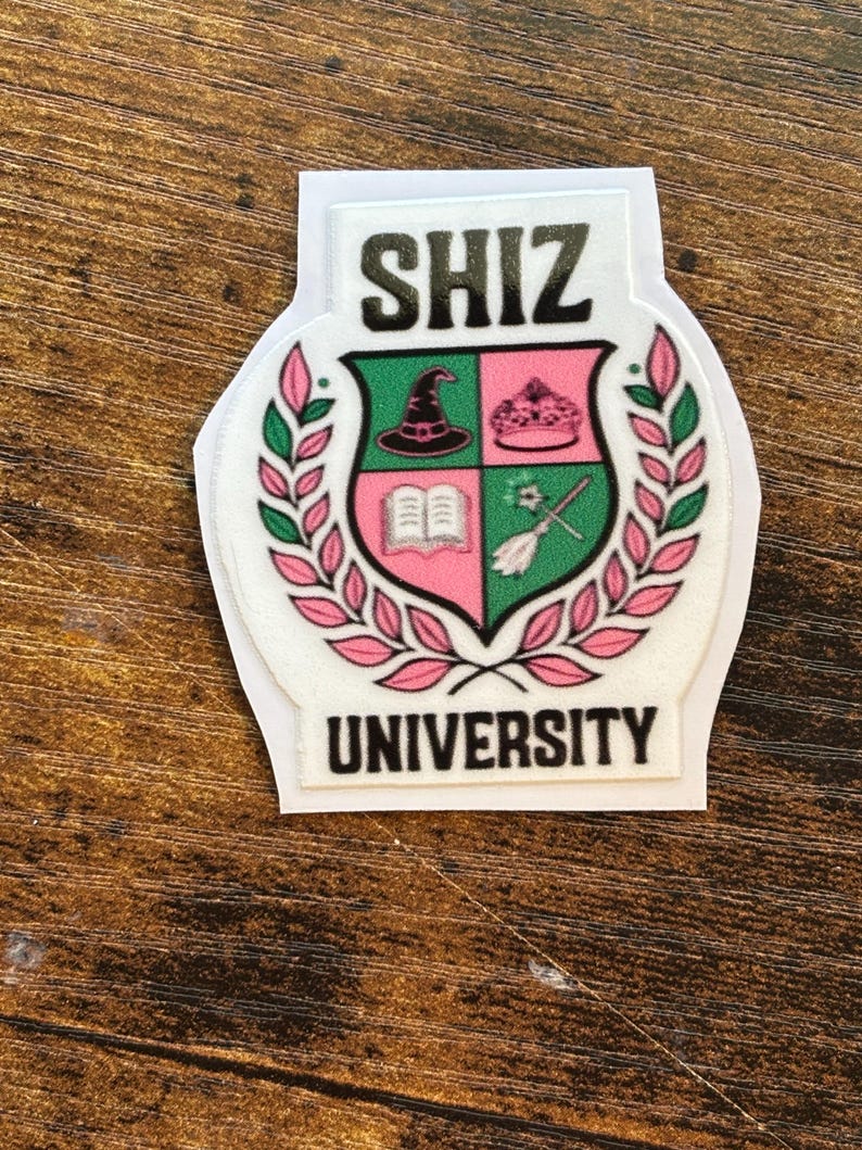 Może przedstawiać: Biała naklejka z czarnym napisem "SHIZ UNIVERSITY". Naklejka przedstawia herb w zielono-r&oacute;żowej kolorystyce, zawierający kapelusz czarownicy, koronę, książkę i miotłę.