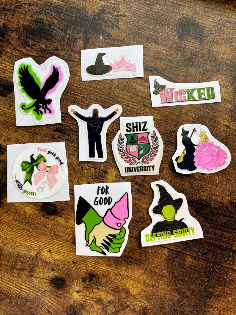 Może przedstawiać: Kolekcja kolorowych naklejek z wzorami i tekstami z musicalu "Wicked". Naklejki zawierają obrazy kapelusza czarownicy, czarnego ptaka oraz słowa "Wicked", "For Good" i "Defying Gravity".