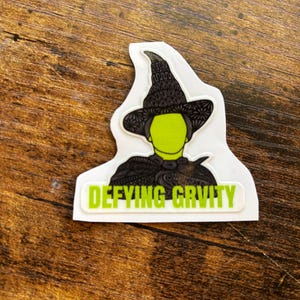 Może przedstawiać: Naklejka przedstawiająca zielonosk&oacute;rą wiedźmę w czarnym kapeluszu i pelerynie. Tekst "Defying Gravity" jest wydrukowany na zielono na białym prostokącie pod wizerunkiem wiedźmy. Naklejka znajduje się na brązowej drewnianej powierzchni.