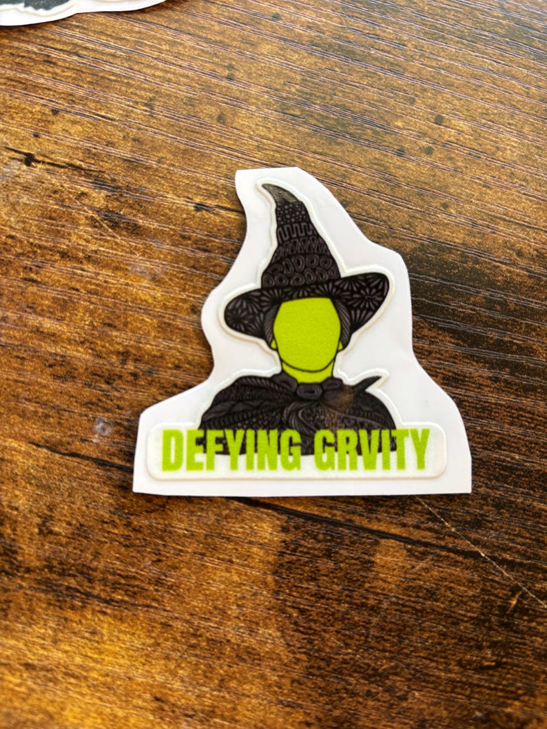 Może przedstawiać: Naklejka przedstawiająca zielonosk&oacute;rą wiedźmę w czarnym kapeluszu i pelerynie. Tekst "Defying Gravity" jest wydrukowany na zielono na białym prostokącie pod wizerunkiem wiedźmy. Naklejka znajduje się na brązowej drewnianej powierzchni.
