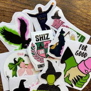 Może przedstawiać: Kolekcja kolorowych naklejek z obrazami i tekstem z musicalu "Wicked". Projekty obejmują sylwetki, kapelusze czarownic i frazy takie jak "Defying Gravity" i "For Good".