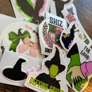 Może przedstawiać: Kolekcja naklejek z r&oacute;żnymi wzorami i tekstem. Wzory obejmują sylwetkę osoby, orła, czarownicy oraz tekst, kt&oacute;ry brzmi "SHIZ", "Pink goes" i "Defying Gravity".