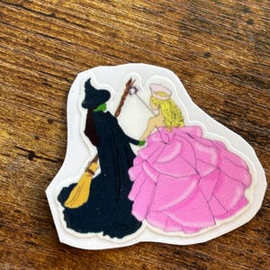 Może przedstawiać: Wycięta naklejka przedstawiająca dwie postacie z Czarnoksiężnika z Oz. Zła Czarownica z Zachodu jest przedstawiona w czerni z zieloną twarzą, trzymając miotłę. Glinda Dobra Czarownica jest w r&oacute;żowej sukni, trzymając r&oacute;żdżkę. Naklejka jest na białym tle.