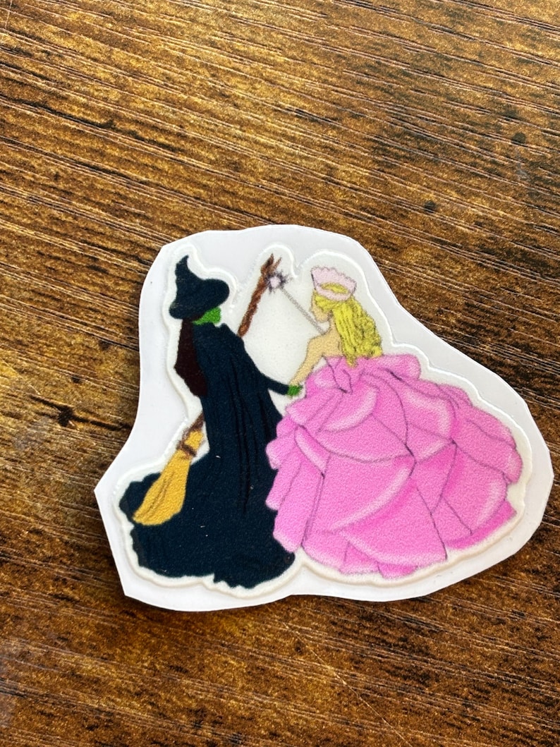 Może przedstawiać: Wycięta naklejka przedstawiająca dwie postacie z Czarnoksiężnika z Oz. Zła Czarownica z Zachodu jest przedstawiona w czerni z zieloną twarzą, trzymając miotłę. Glinda Dobra Czarownica jest w r&oacute;żowej sukni, trzymając r&oacute;żdżkę. Naklejka jest na białym tle.