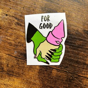 Może przedstawiać: Naklejka przedstawiająca dwie złączone dłonie z kresk&oacute;wek. Jedna dłoń jest zielona z czarnymi paznokciami, druga ma jasny odcień sk&oacute;ry. Dłonie połączone są r&oacute;żowym mankietem. Nad dłońmi wydrukowano słowa "FOR GOOD".