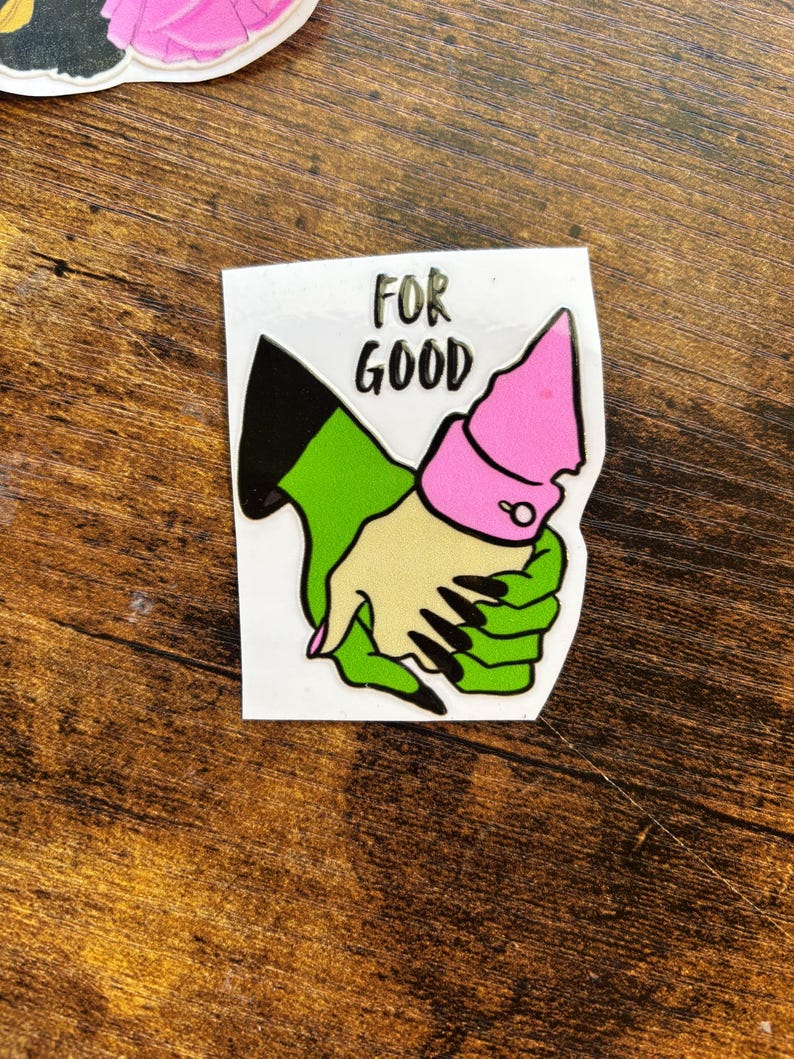 Może przedstawiać: Naklejka przedstawiająca dwie złączone dłonie z kresk&oacute;wek. Jedna dłoń jest zielona z czarnymi paznokciami, druga ma jasny odcień sk&oacute;ry. Dłonie połączone są r&oacute;żowym mankietem. Nad dłońmi wydrukowano słowa "FOR GOOD".