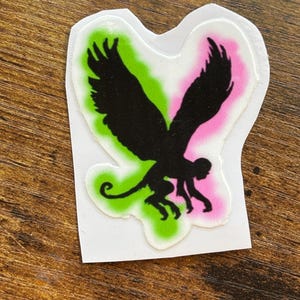 Może przedstawiać: Wycięta naklejka z czarną sylwetką hybrydy małpy i ptaka. Projekt umieszczono na zielono-r&oacute;żowym tle gradientowym z białym obramowaniem. Na drewnianej powierzchni.