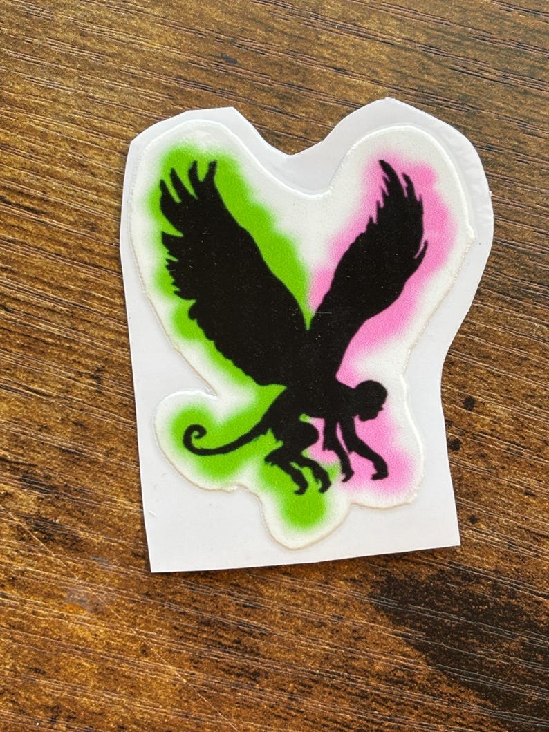 Może przedstawiać: Wycięta naklejka z czarną sylwetką hybrydy małpy i ptaka. Projekt umieszczono na zielono-r&oacute;żowym tle gradientowym z białym obramowaniem. Na drewnianej powierzchni.