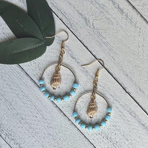 Peut inclure: Paire de boucles d'oreilles dorées avec un charme en forme de coquillage et des perles bleues claires et dorées. Les boucles d'oreilles sont présentées sur une surface à grain de bois clair.