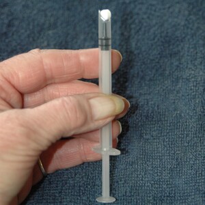 Peut inclure: Une seringue en plastique transparente avec une pointe blanche et un anneau noir autour du piston. La seringue est tenue dans une main.