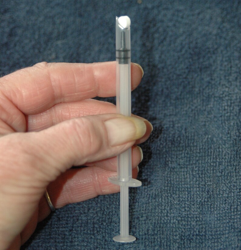 Peut inclure: Une seringue en plastique transparente avec une pointe blanche et un anneau noir autour du piston. La seringue est tenue dans une main.