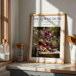May include: A framed print featuring the text "THE QUR'AN (26:79)" and a quote. The artwork shows a still life of food and flowers. The print is in a light wood frame and is displayed on a countertop near a window.