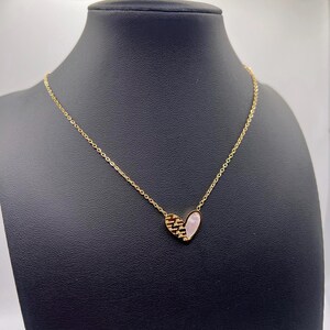 Könnte beinhalten: Eine goldene Kettenkette mit einem herzförmigen Anhänger. Der Anhänger besteht aus einer rosa Muschel mit einem goldenen Design auf einer Seite.
