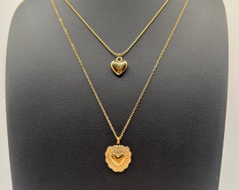 May include: A gold layered necklace with two heart pendants. The top pendant is a simple heart shape, while the bottom pendant is a more ornate heart with a raised design.