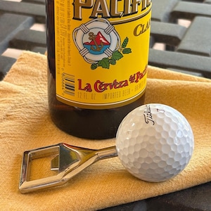 May include: A silver metal bottle opener with a white Titleist golf ball handle positioned in front of a brown glass Pacifico beer bottle. The bottle features a yellow label with a life preserver and anchor graphic and the text La Cerveza del Pacifico.