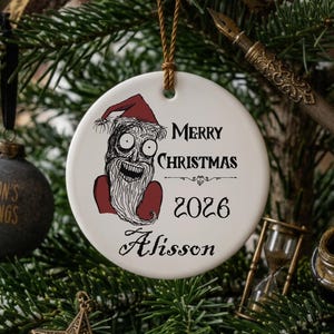 Puede incluir: Un adorno redondo de cerámica blanca con una ilustración estilizada de una figura barbuda con un gorro de Papá Noel rojo. El texto dice Merry Christmas, 2026, y el nombre Alisson en letra negra. Incluye un cordel marrón para colgarlo.
