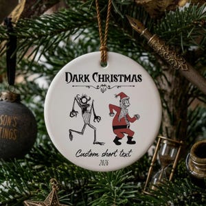 Puede incluir: Un adorno redondo de cerámica blanca con el texto negro Dark Christmas, un dibujo lineal de un monstruo con cuernos y una figura de Papá Noel en rojo y blanco. Debajo de las figuras, el texto dice Custom short text y el año 2026 en negro.